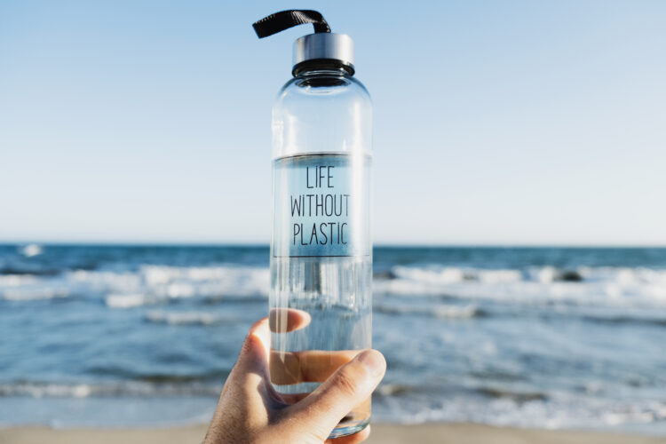 closeup of a man holding a glass reusable water bottle with the text life without plastic written in it, on the beach, with the ocean in the background - earth day at home